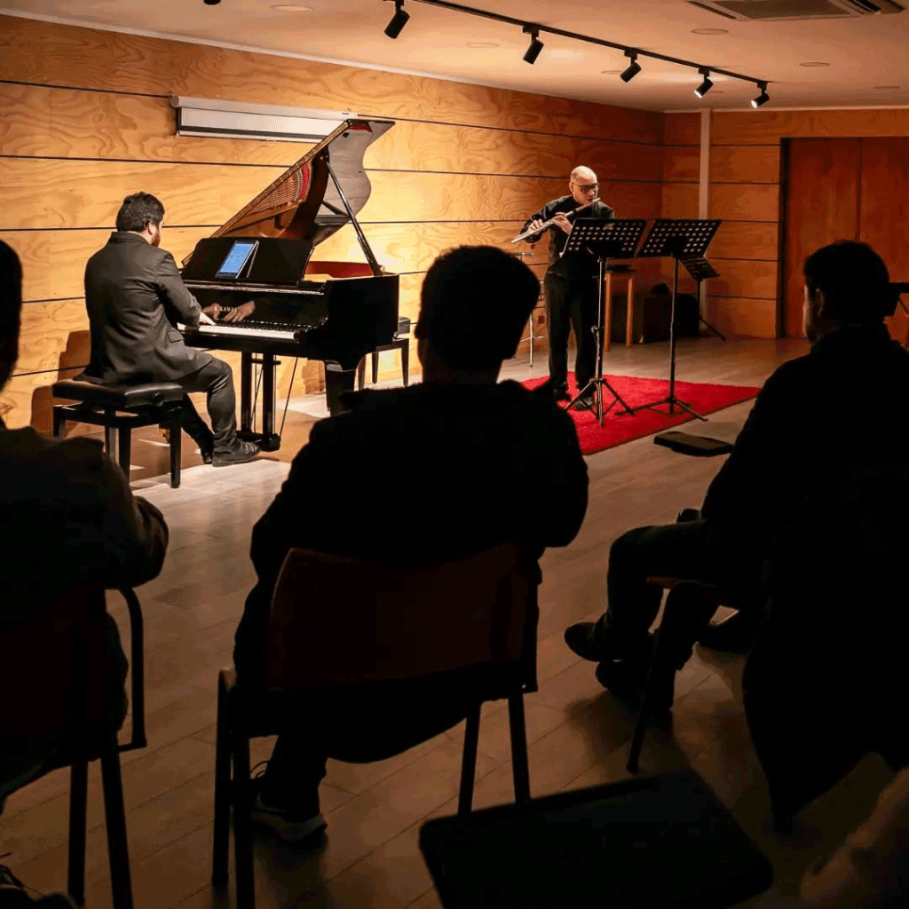 Imagen de siluetas de personas con el dúo en piano y flauta, tocando de fondo.